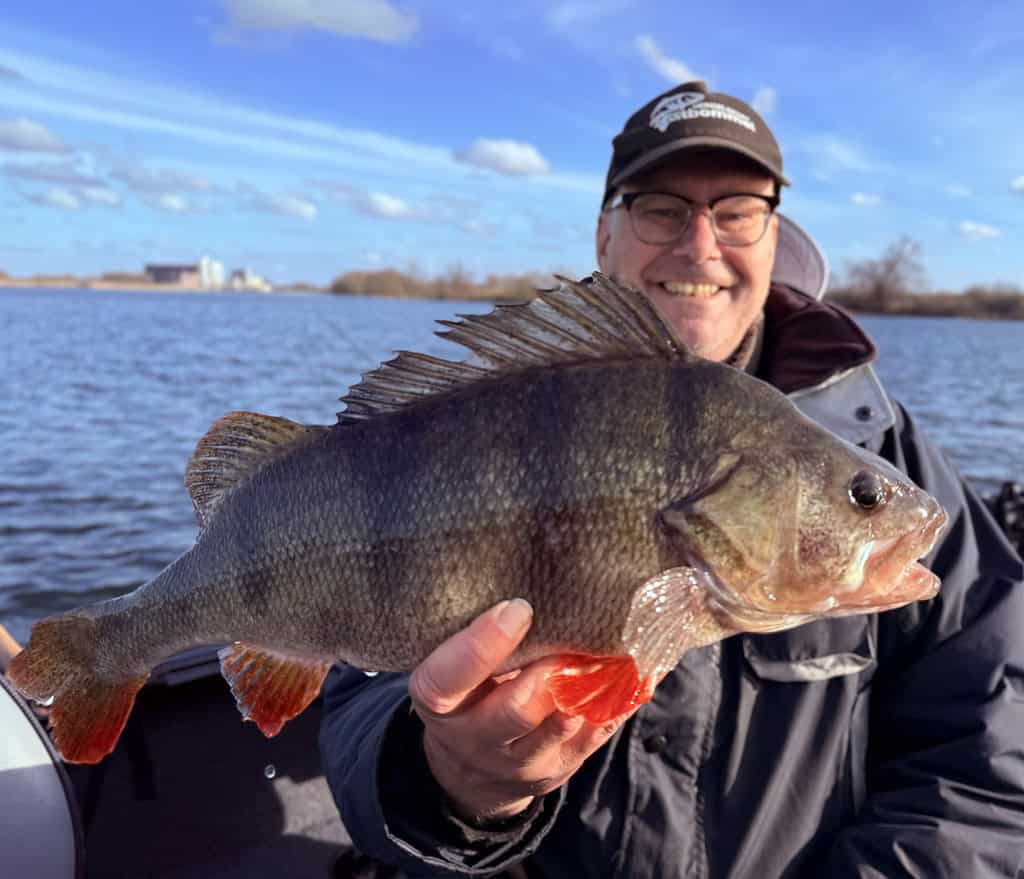 Een persoon met een jas, pet en bril lacht breed terwijl hij trots een grote vis vasthoudt op zijn boot. De serene achtergrond van het meer vult de gedeeltelijk bewolkte lucht aan terwijl ze hun indrukwekkende vangsten laten zien met behulp van de vertrouwde Predatorgear.