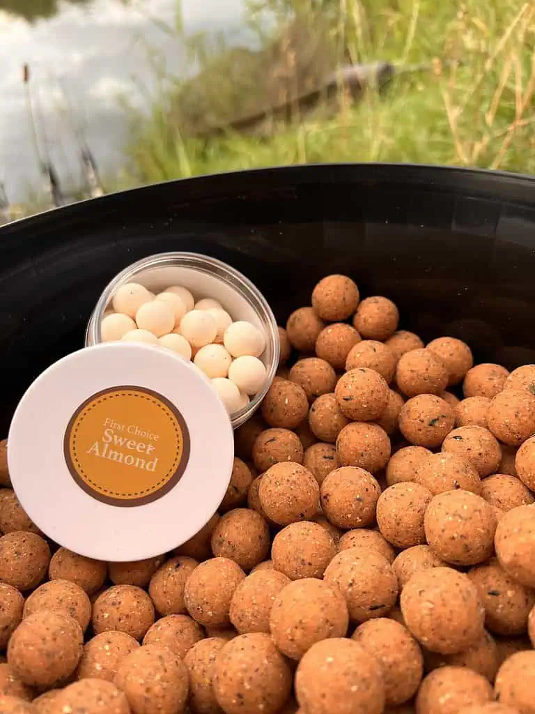 Een kom met bruine aasballen staat naast een vijver, vergezeld door een klein open bakje met crèmekleurig "First Choice Sweet Almond" aas. Perfect voor die lente-karpervisavonturen, deze zijn essentieel voor de verzameling tips & trucs van elke visser.