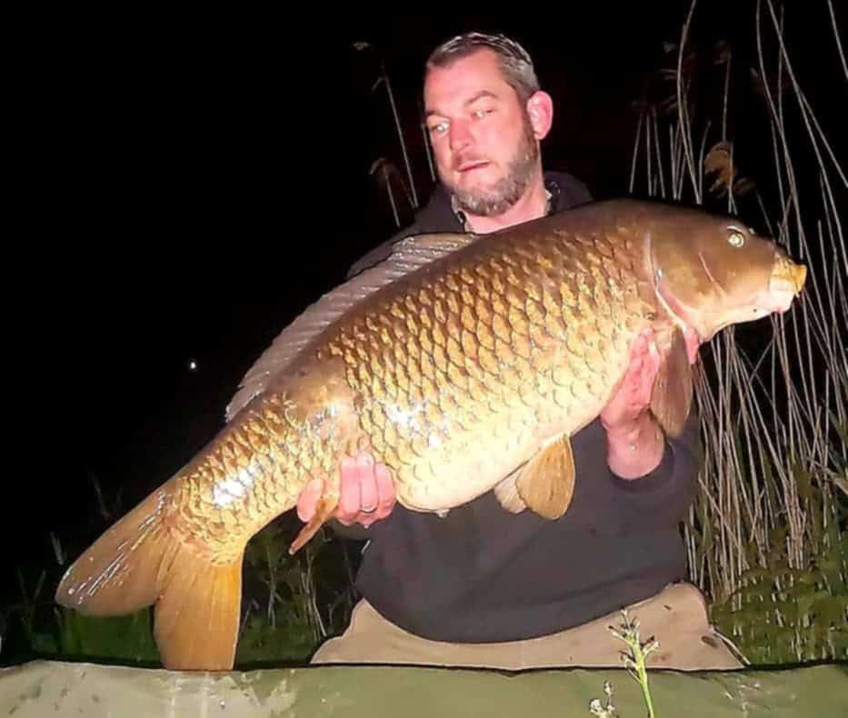 Onder de maanverlichte hemel houdt een man trots een grote vis vast, gevangen tijdens een avontuurlijke nacht karpervissen, midden in het fluisterende hoge gras.