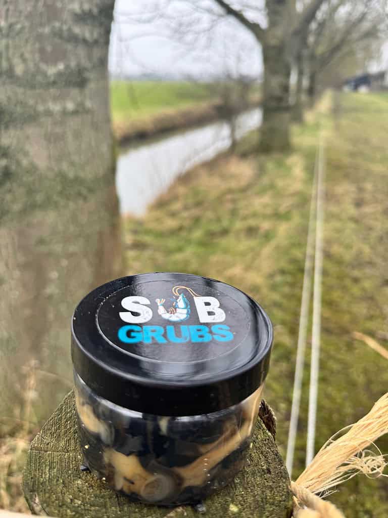 Een klein potje met het opschrift "Sub Grubs" staat op een houten paal, vlakbij een weelderig grasveld vol bomen en een rustig kanaal op de achtergrond. Ideaal om penvissen aan te trekken.