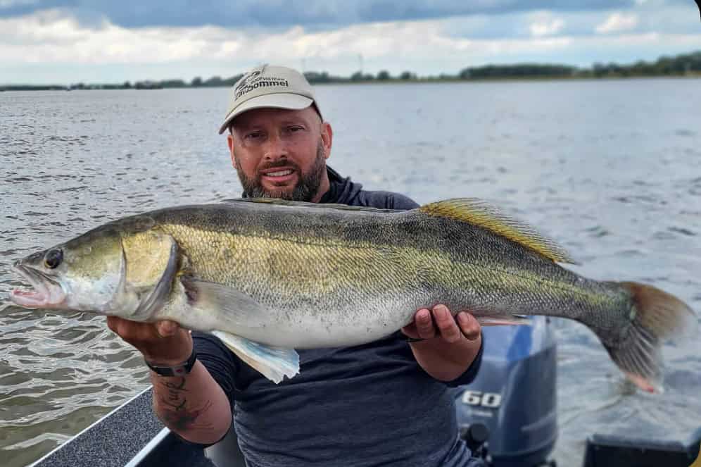 Een persoon met een hoed op houdt een grote vis vast terwijl hij in een boot op het water zit en zijn volgende avontuur plant op basis van de agenda van Hengelsport Zaltbommel Open Dagen.