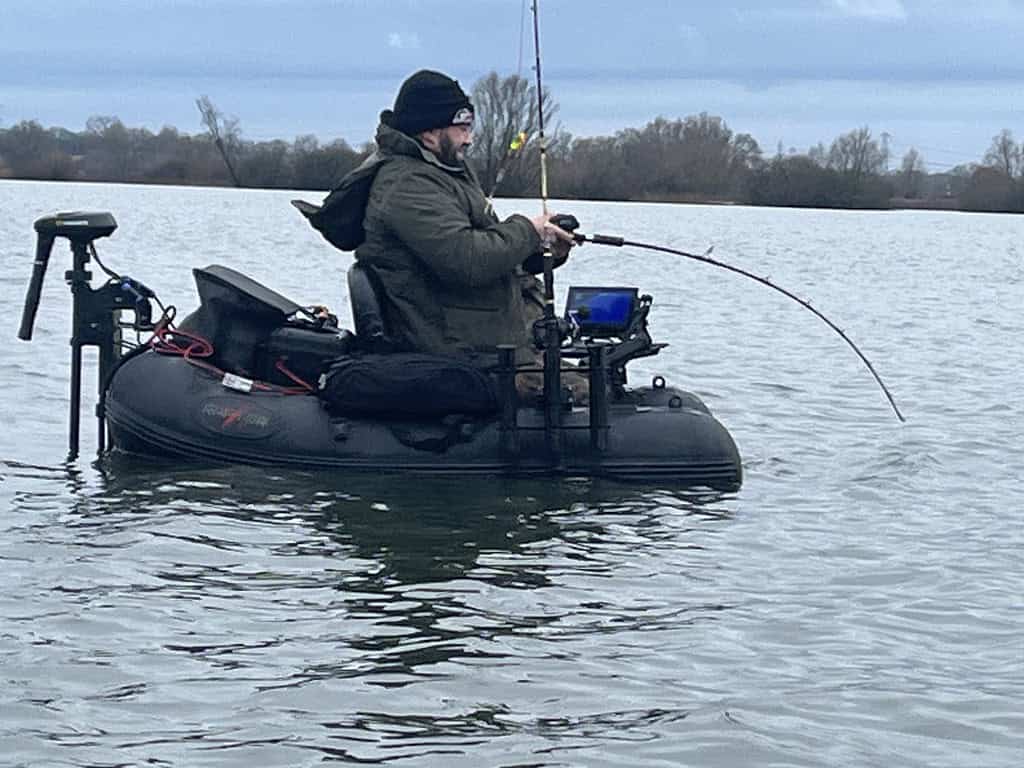 Een man in een zwarte opblaasbare bellyboat vist op een meer onder een bewolkte hemel. Hij draagt een groen jack en een zwarte muts.