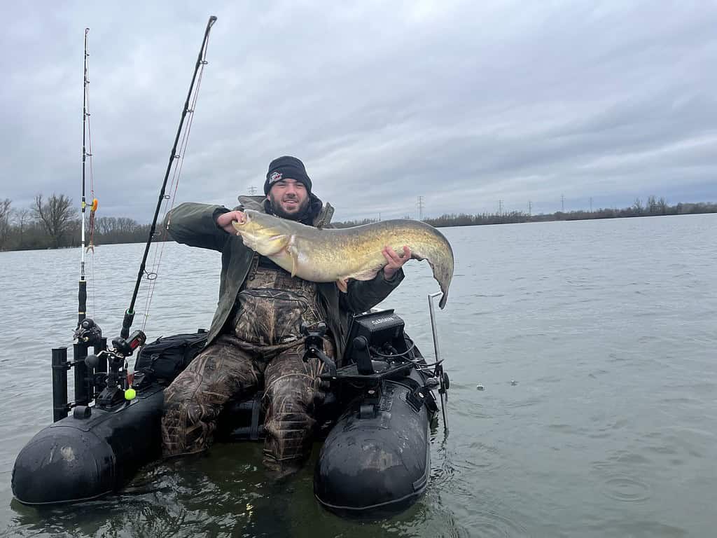 Een persoon in een camouflagepak zit in een opblaasbare bellyboat en houdt trots een monsterlijke vis vast. Naast zich liggen hengels op een bewolkte dag op het meer.