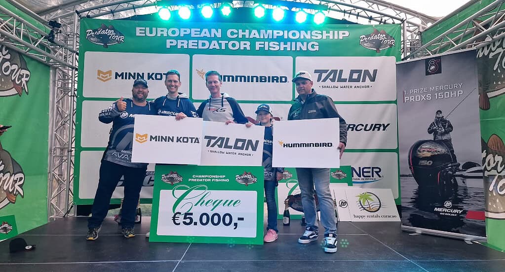 Een groep van vijf mensen houdt trots sponsorborden en een cheque van € 5.000 vast op het podium van het Europees Kampioenschap Roofvissen, met mediapartner Viswereld en Predatortour op de achtergrond.