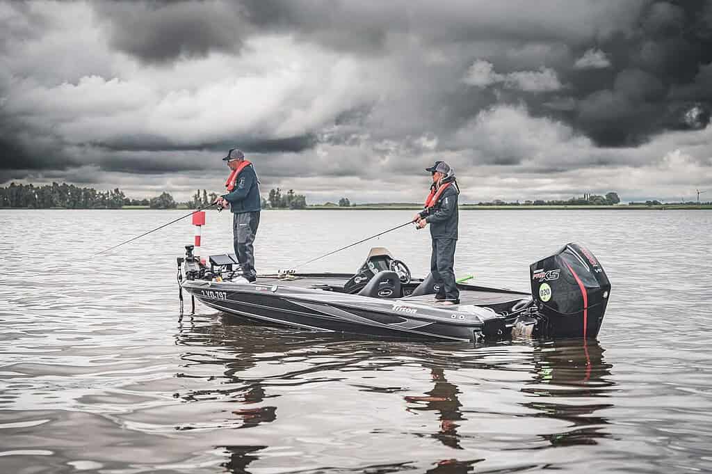 Twee mensen vissen op een boot in een groot water, deelnemend aan de beroemde Predatortour. Beiden dragen hoeden en zwemvesten terwijl ze hun lijnen uitwerpen onder een bewolkte, bewolkte lucht.