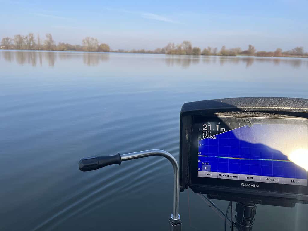 Een kalm meer met een dieptemeterdisplay op een boot, die 21,1 meter diepte aangeeft, ideaal om meerval te spotten. Bomen zijn zichtbaar op de achtergrond onder een heldere hemel, waardoor het een sereen tafereel is, perfect voor liefhebbers van wintervisserij.