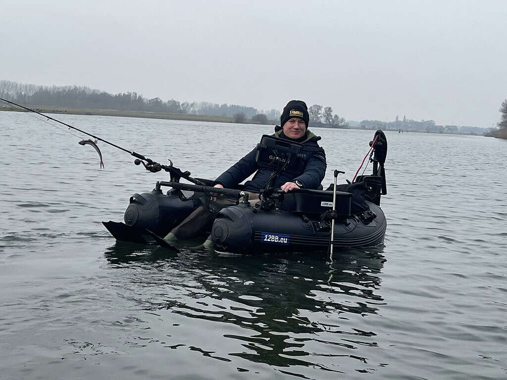 Persoon in een kleine opblaasbare boot op een meer, met een zwemvest en een hoed, en een hengel in de hand. Bewolkte lucht en verre boomgrens op de achtergrond.