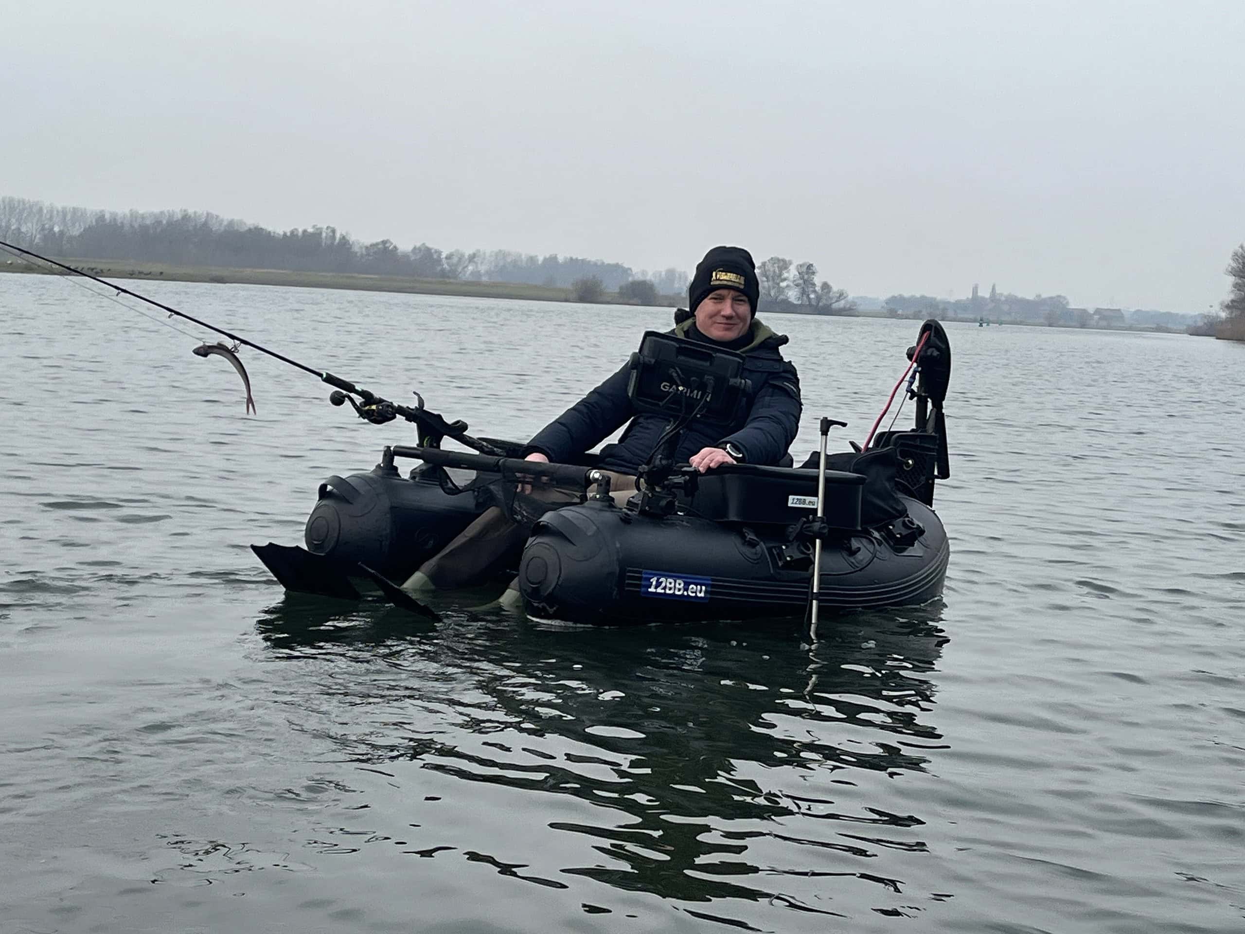 Persoon in een zwart jasje en hoed die vist vanaf een kleine opblaasbare boot op een kalm meer. Bewolkte lucht en een kustlijn met bomen op de achtergrond.