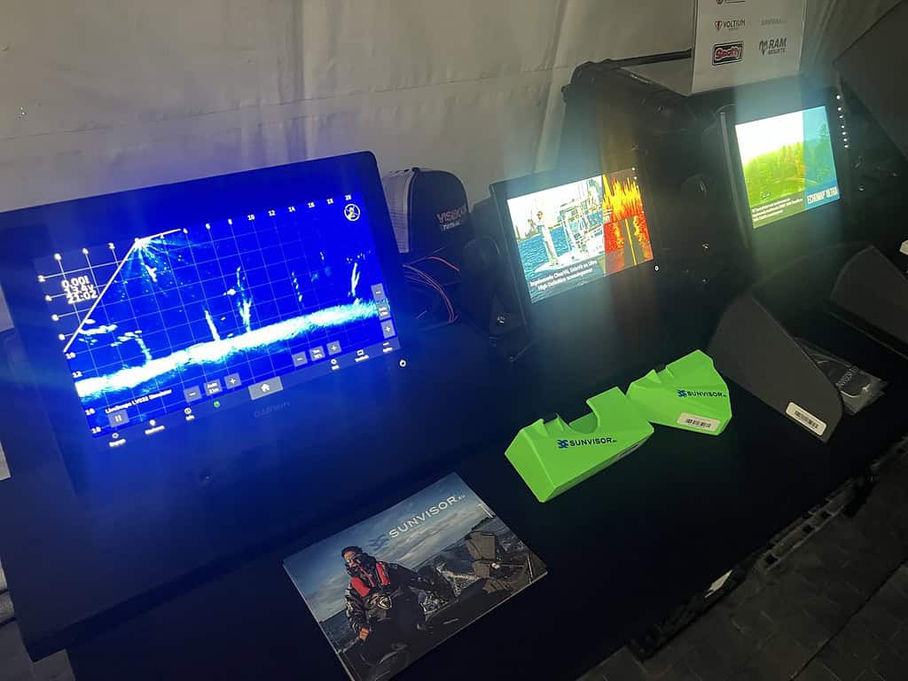 Een display van elektronische apparaten siert de tafel, met schermen met diverse data, omringd door brochures en merkpetten van Hengelsport Zaltbommel. Ontdek het tech-aanbod op onze open dagen in Bommelerwaard.