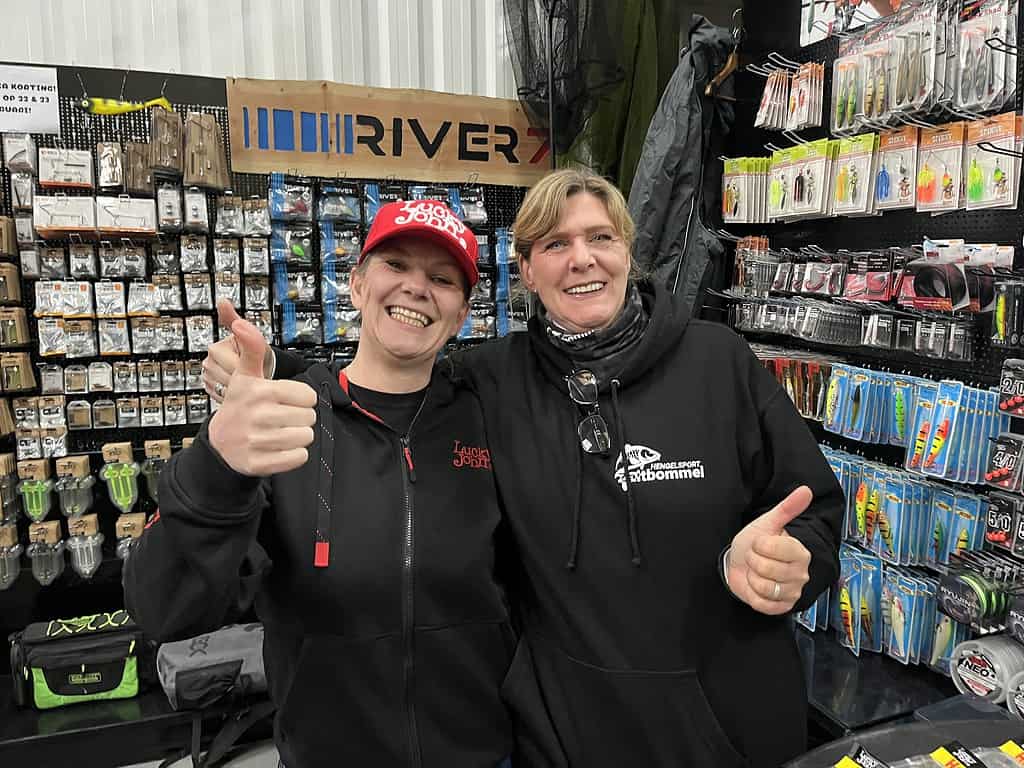 Twee mensen steken hun duim omhoog bij Hengelsport Zaltbommel tijdens de open dagen, omringd door rekken vol verpakte hengelsportspullen in de gezellige winkel in de Bommelerwaard.