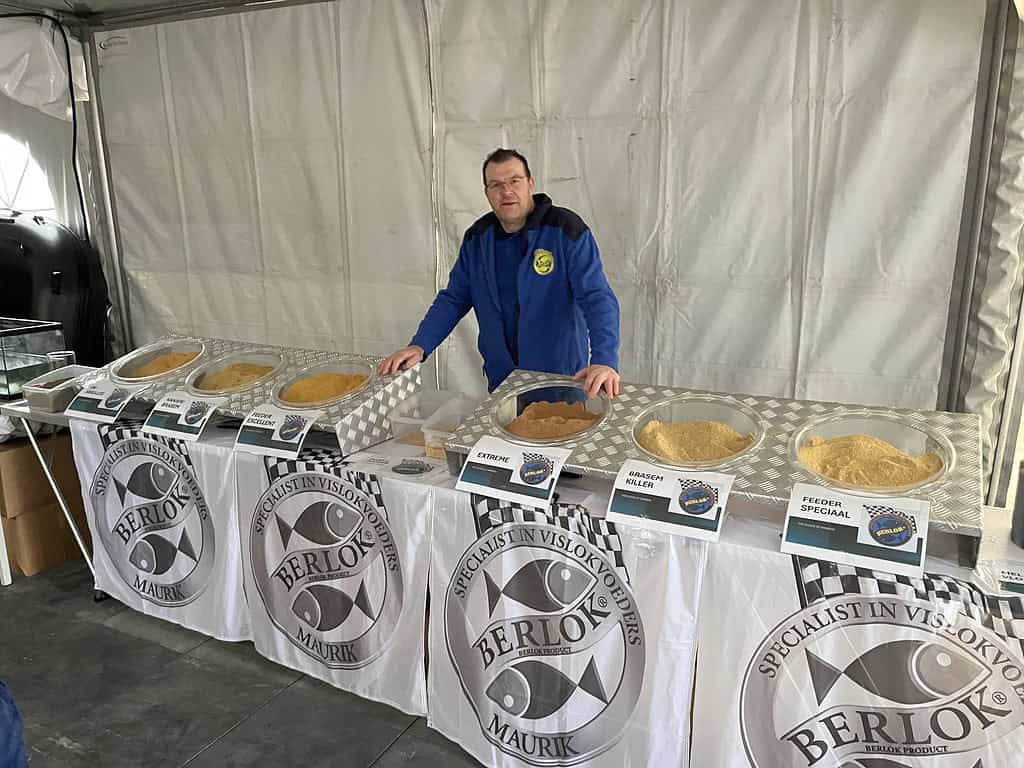 Een persoon staat achter een tafel met bakjes visvoer, elk gelabeld met verschillende soorten. Een banner met de tekst "Berlin" en een logo zijn zichtbaar op het tafelkleed, wat doet denken aan een levendig Hengelsport-evenement in Zaltbommel.