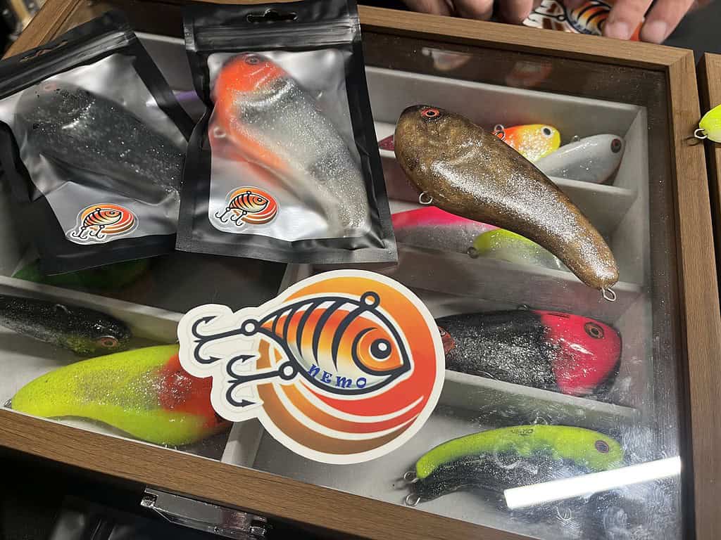 Een vitrine toont verschillende kleurrijke vislokmiddelen, sommige nog in de verpakking, compleet met een "Nemo"-sticker met een abstract oranje visontwerp. Perfect voor elke Hengelsportliefhebber die de levendige aanbiedingen van Bommelerwaard of Zaltbommel verkent.