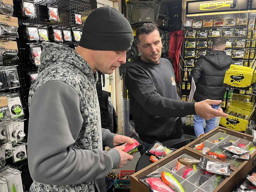 Twee mensen in een winkel bekijken vislokmiddelen die op een tafel zijn uitgestald, terwijl planken vol met verschillende visuitrustingen en accessoires de achtergrond vormen. Het is een waar visfeestje bij Hengelsport Zaltbommel, perfect voor liefhebbers van open dagen die op zoek zijn naar het beste op het gebied van hengelsportuitrusting.