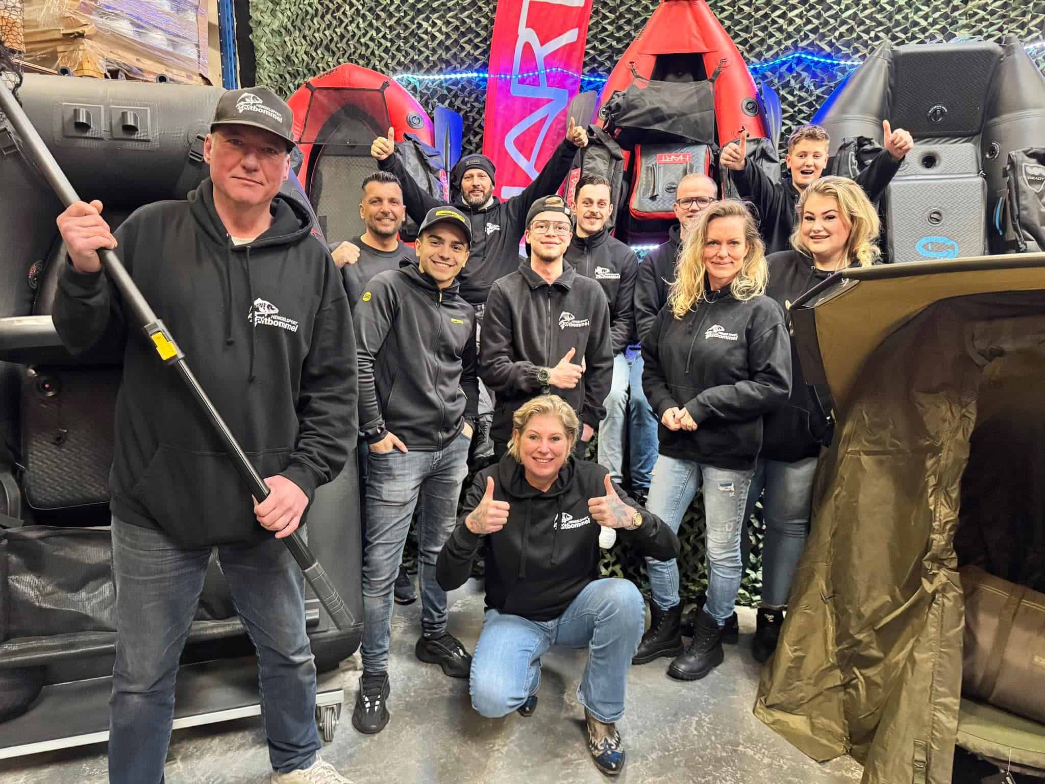 Een groep van elf mensen die binnen poseren, omringd door opblaasboten en kampeeruitrusting, legt de essentie van de Bommelerwaard-spirit vast. Meerdere dragen zwarte hoodies met een logo. Een vrouw knielt vooraan en steekt haar duim op, wat de kameraadschap belichaamt die je vindt in Hengelsport-avonturen.