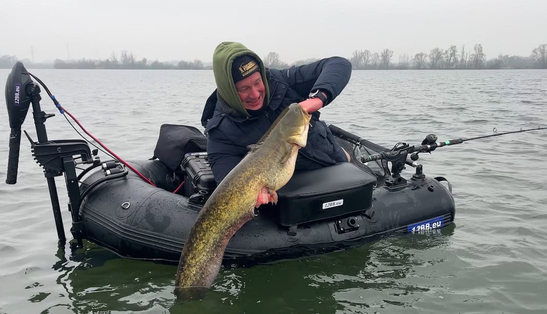 Een persoon zit in een kleine opblaasbare boot op het water, trots een grote vangsten in beide handen vasthoudend, omringd door visgerei. Bomen vormen een serene achtergrond voor deze succesvolle week op het meer.