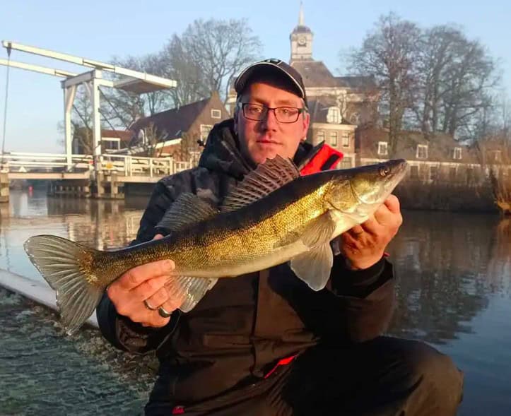 Een persoon met een Predatorgear-bril en een pet houdt trots een grote vis vast voor een schilderachtig uitzicht op het water. Hier zou "Vangsten van de maand" gemakkelijk gevierd kunnen worden, met de brug en het gebouw als perfecte achtergrond.