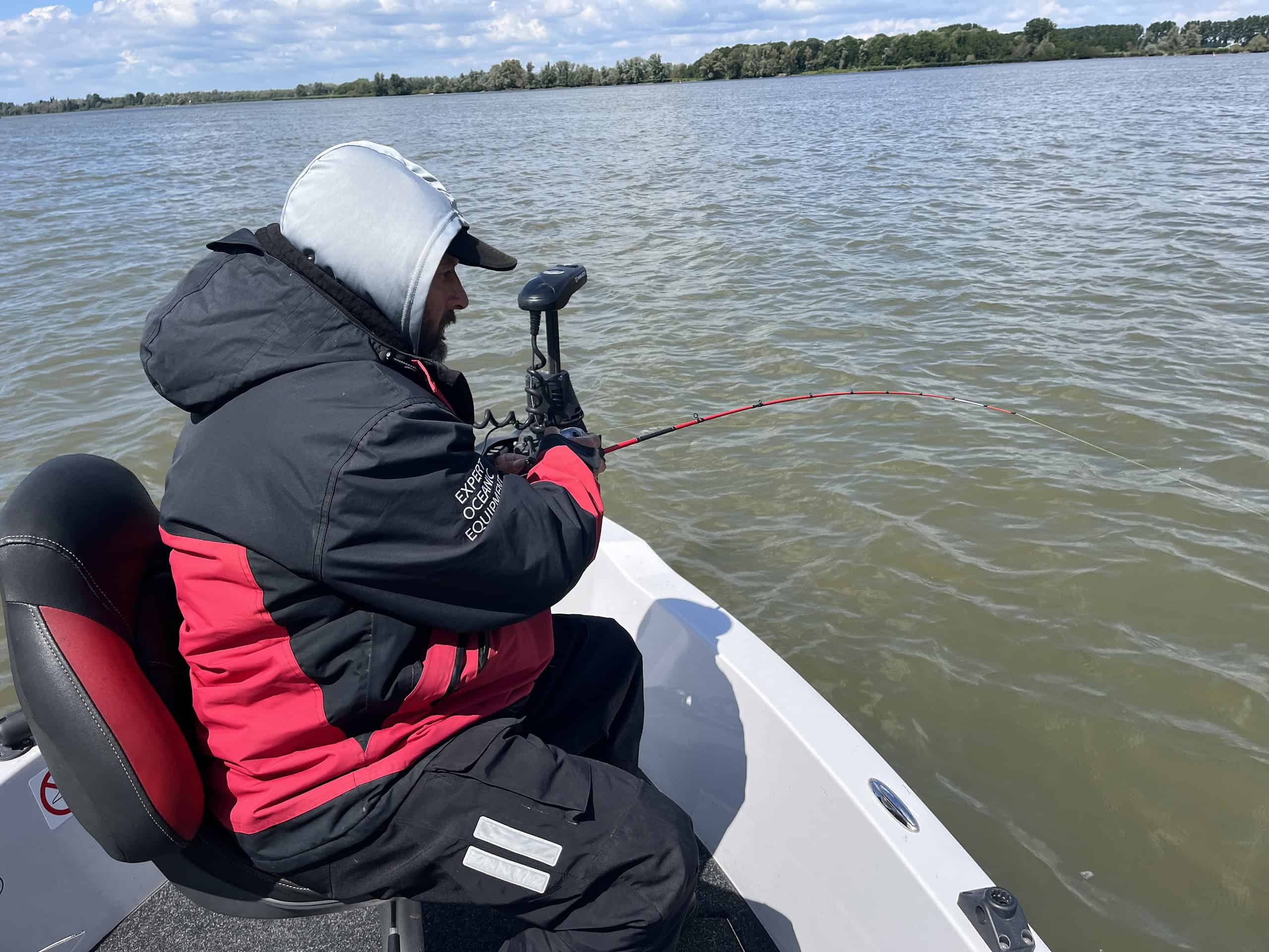 Persoon in een rood en zwart jasje, zittend op een bootstoel terwijl hij op een bewolkte dag in een grote watermassa voorvissen.