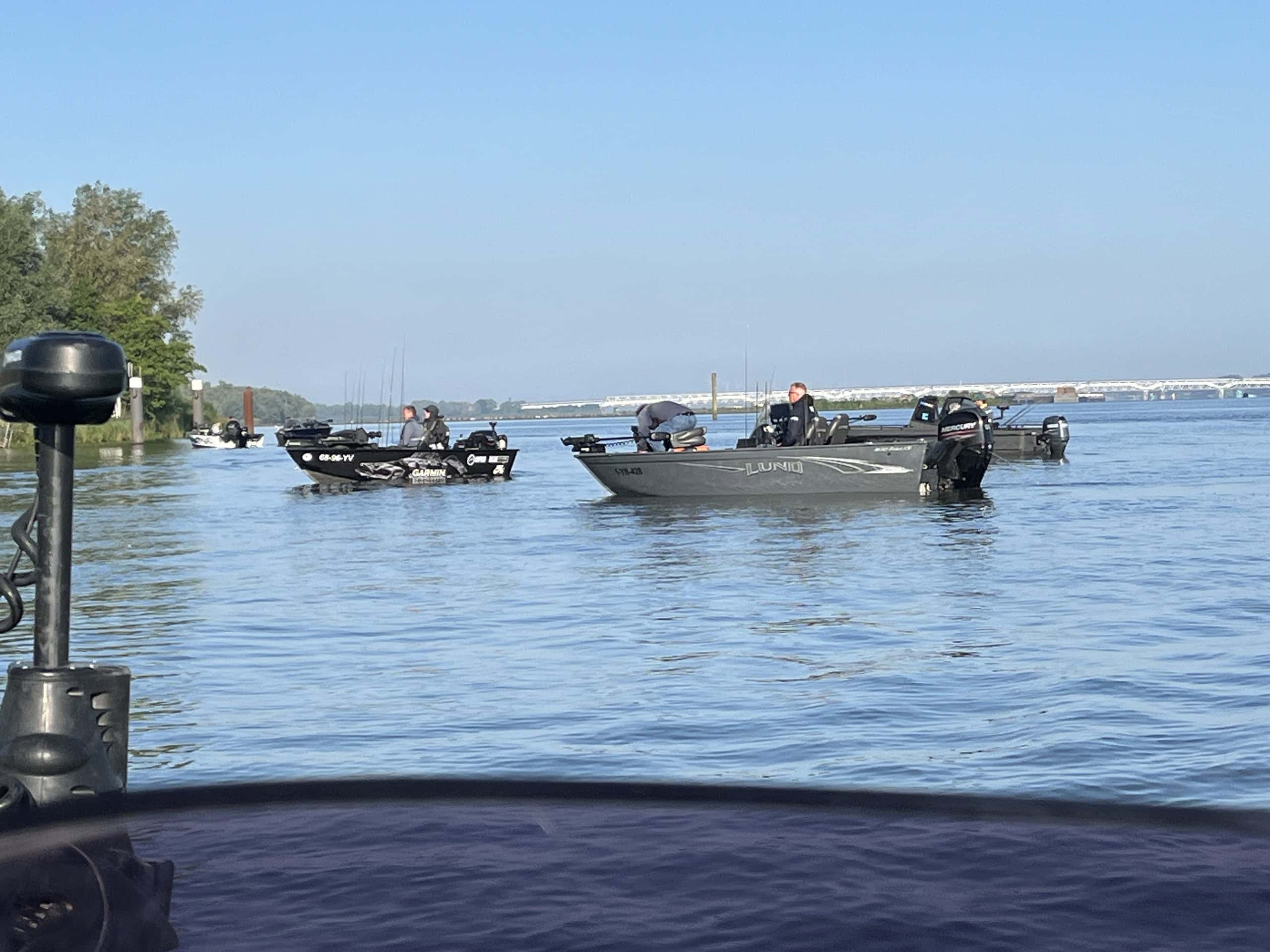 Op een kalm water liggen verschillende kleine motorboten, elk met individuen die zich bezighouden met visserijactiviteiten. Onder een strakblauwe hemel staan bomen langs de verre oever, terwijl de deelnemers gretig meedoen aan de dakviswedstrijd.