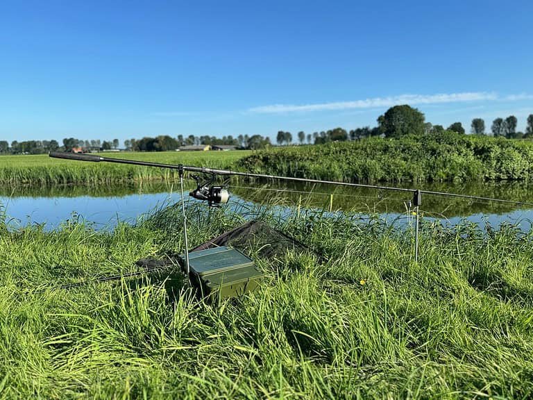 Hengel uitgezet op een grazige oever bij een kalme rivier met groene velden en bomen op de achtergrond onder een helderblauwe lucht, perfect voor een rustig dagje polderkarpervissen.