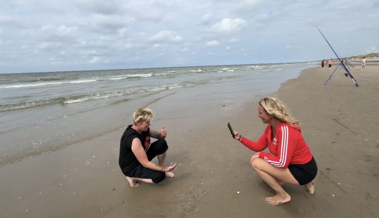 Twee mensen staan op een zandstrand. Een persoon hurkt en steekt zijn duim omhoog, terwijl een ander knielt om een foto te maken met een smartphone. Op de achtergrond staat een hengel opgesteld, die de essentie van strandvissen vastlegt.