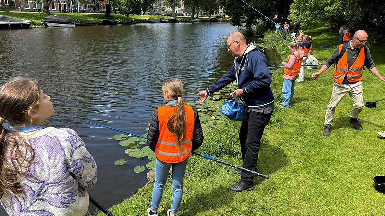 Een groep mensen, bestaande uit kinderen en volwassenen, draagt oranje hesjes en is aan het vissen aan de oever van een kalme rivier op een grasveld in een parkachtige omgeving. Ze vangen de essentie van conceptvorming te midden van de rust van de natuur.