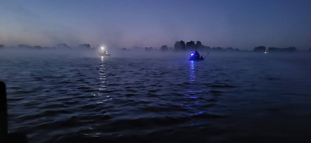 Twee boten met blauwe lichten drijven op een mistig water in de schemering, wat een griezelig mooi tafereel creëert. Silhouetten van bomen langs de horizon, wat bijdraagt aan de mysterieuze sfeer. Het is alsof de scène uit een spannend deel 5 van een boeiende serie komt.