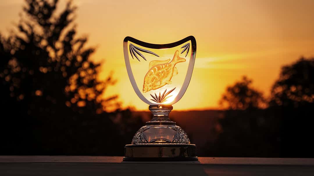 Een glazen trofee met de afbeelding van een vis wordt belicht door een zonsondergang boven het Lac de Madine, met het silhouet van bomen op de achtergrond.