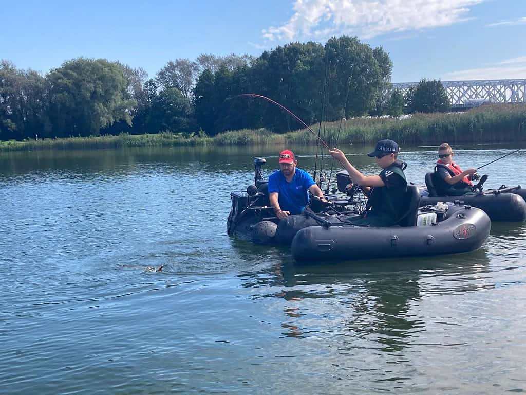 Drie mensen zijn aan het sportvissen in een kleine opblaasbare boot op een kalm meer met bomen en een brug op de achtergrond. Eén persoon is actief bezig met het binnenhalen van een vis, wat de spanning van de bellyboat wedstrijd laat zien.