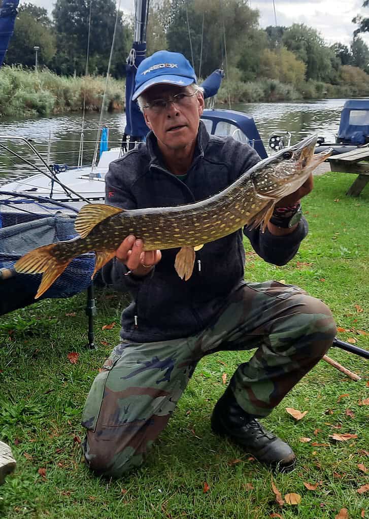 Een persoon met een blauwe pet en een camouflagebroek, mogelijk Krijn van Urk na een succesvolle doodaassessie, houdt een grote vis vast bij een rivier met op de achtergrond aangemeerde boten.