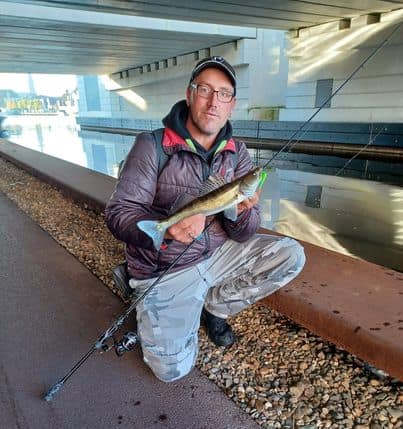 Een persoon knielt bij een kanaal, met een hengel in de ene hand en hun Vangsten in de andere, onder een brug. Ze zijn gekleed in casual outdoorkleding, wat aangeeft dat het najaarsvisseizoen is begonnen.