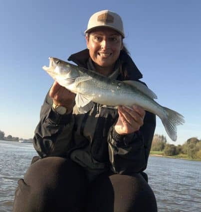 Een persoon met een jas en een pet houdt op een zonnige dag een vis vast aan het water, terwijl hij trots zijn'vangsten van de week' laat zien.