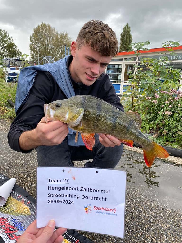 Een persoon in een donker jasje houdt een vis vast, terwijl een ander persoon een kaartje toont met de tekst "Team 27 Hengelsport Zaltbommel Streetfishing Dordrecht 28/09/2024", waarmee hij/zij deelneemt aan de NK Streetfishing kwalificatiewedstrijd.