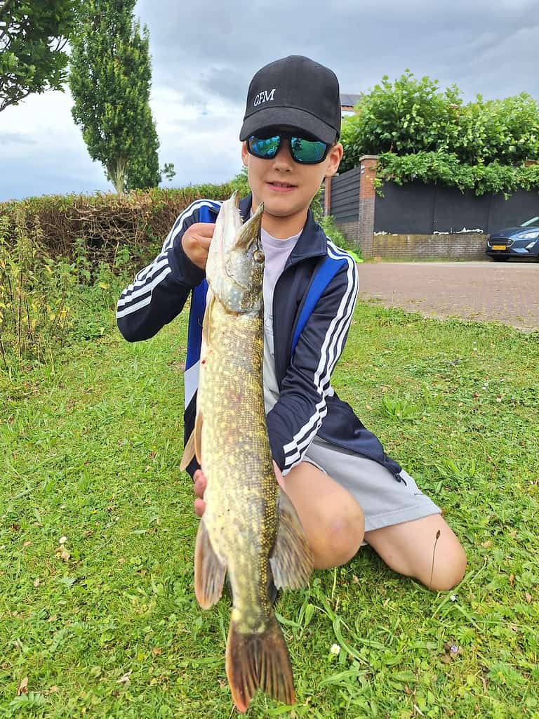 Een kind met een pet en zonnebril knielt op het gras terwijl hij trots een grote vis met beide handen omhoog houdt, en een van hun mooie vangsten laat zien. De achtergrond toont een heg, een boom en een deel van een straat met geparkeerde auto's.