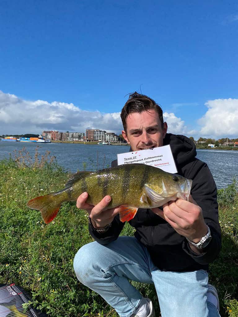 Een persoon met een grote vis en een bord met tekst, staat buiten bij een waterpartij met gebouwen op de achtergrond tijdens de NK Streetfishing kwalificatiewedstrijd in Dordrecht.