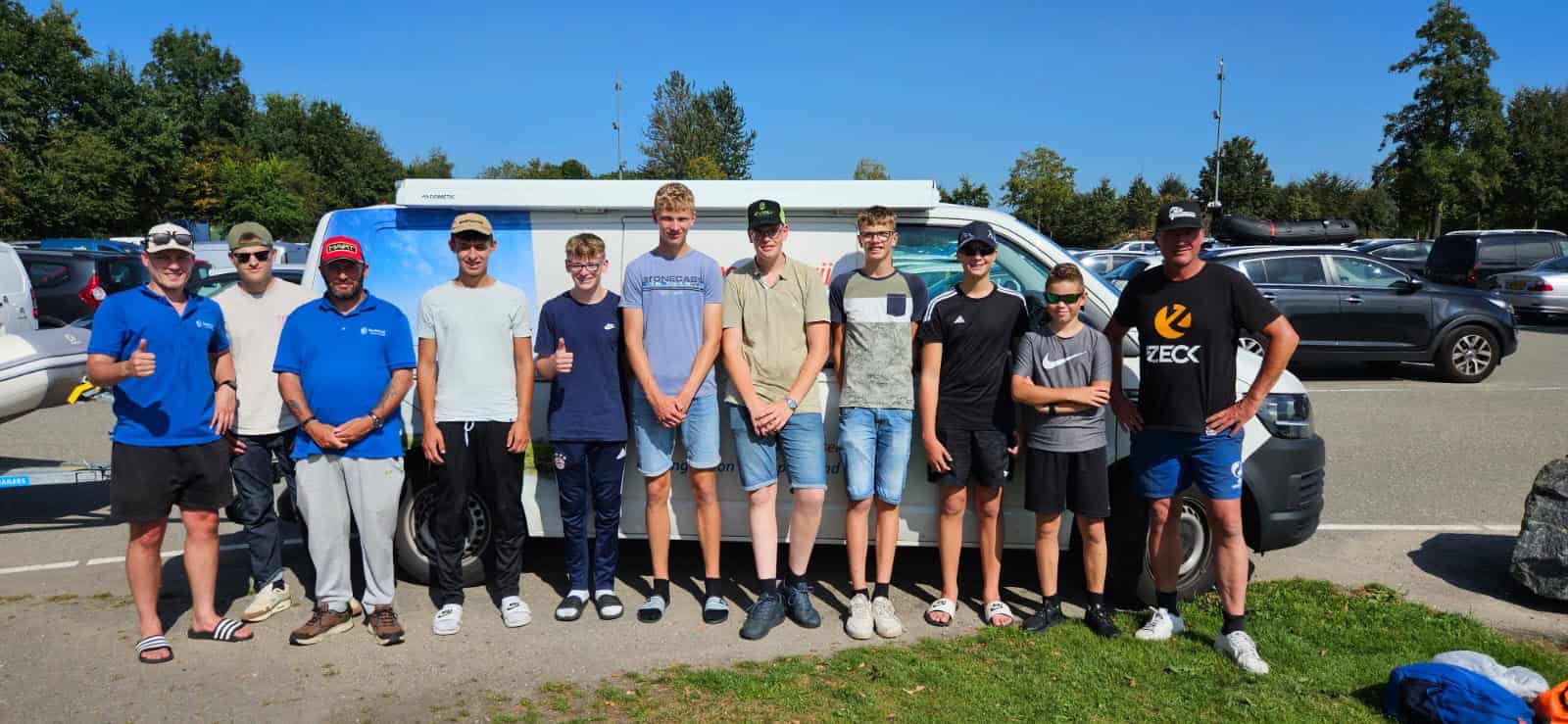 Een groep van elf mensen in casual kleding staat voor een wit busje op een parkeerplaats, waarbij sommigen hun duim omhoog steken. Het tafereel, omlijst door bomen en andere geparkeerde auto's, weerspiegelt het gemak en de efficiëntie die vaak geassocieerd worden met automatische concepten.