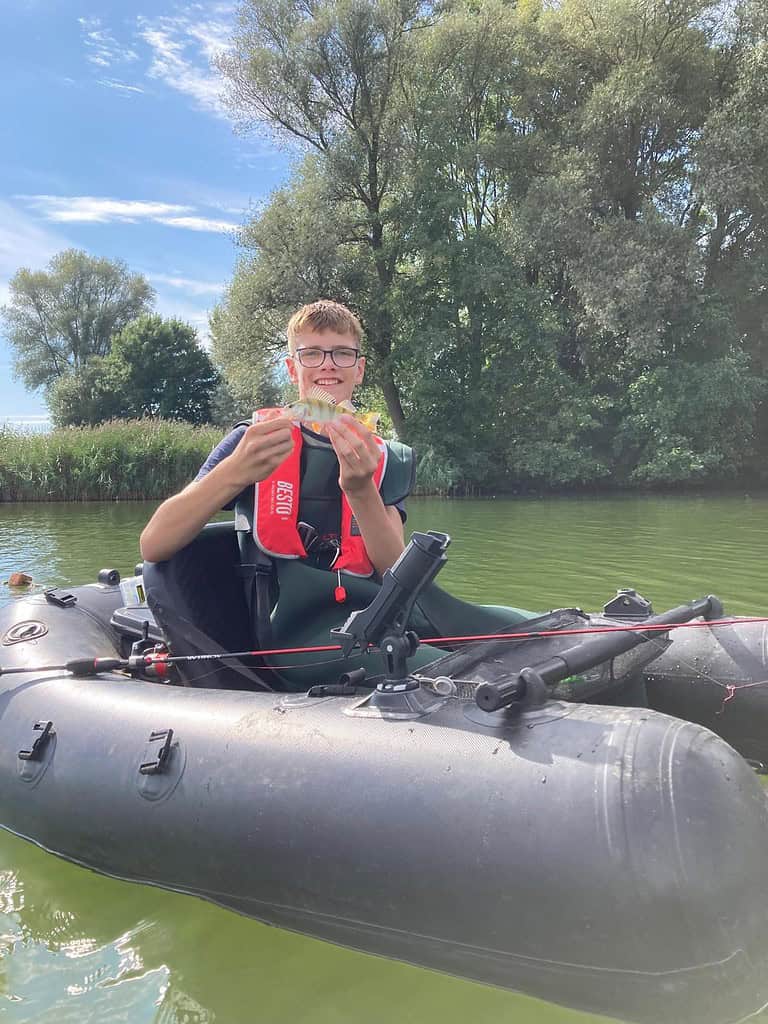 Een persoon zit in een bellyboat met een kleine vis in zijn armen. Hij draagt een zwemvest en een bril. De achtergrond bestaat uit bomen en kalm water.