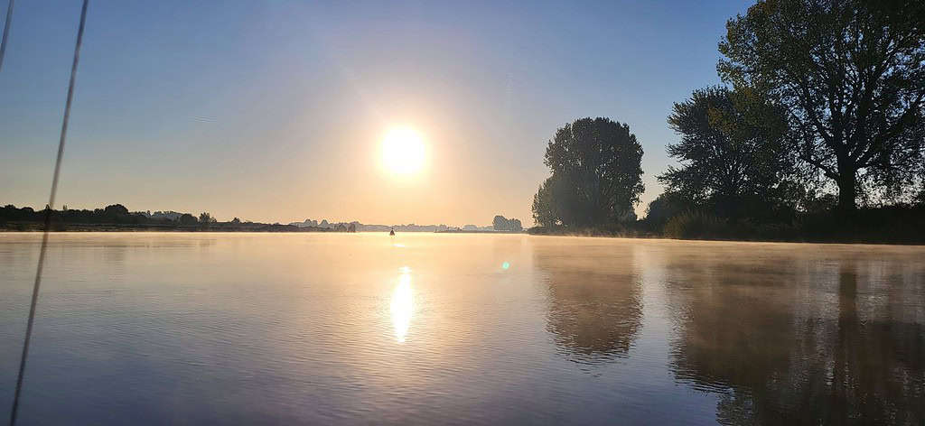 Een rustig meer bij zonsopgang, met mist die opstijgt van het wateroppervlak, grote silhouetten van bomen aan de rechterkant en een heldere, heldere lucht erboven: dit alles zorgt voor een uitzicht dat alle frustratie wegneemt.