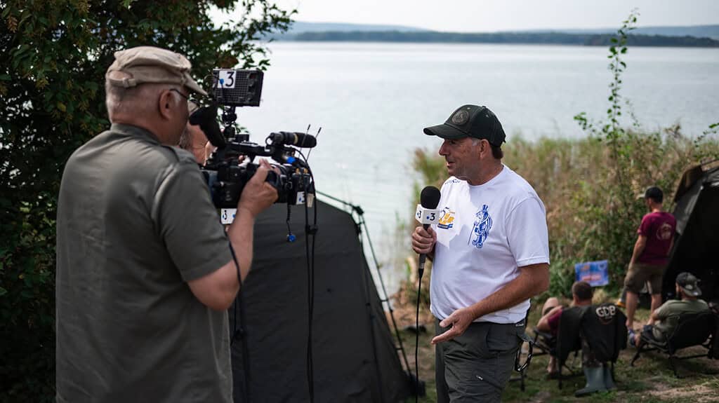 Een man in een wit T-shirt wordt geïnterviewd door een cameraman bij Lac de Madine. De interviewer, die de World Carp Classic 2024 verslaat, houdt een microfoon vast. Andere mensen zitten op de achtergrond bij het water.