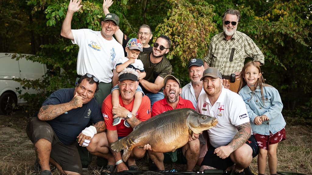 Een groep mensen verzamelt zich lachend rond een persoon die een grote vis vasthoudt en trots poseert tijdens de De World Carp Classic 2024. Bomen en struiken vormen de pittoreske achtergrond van Lac de Madine.