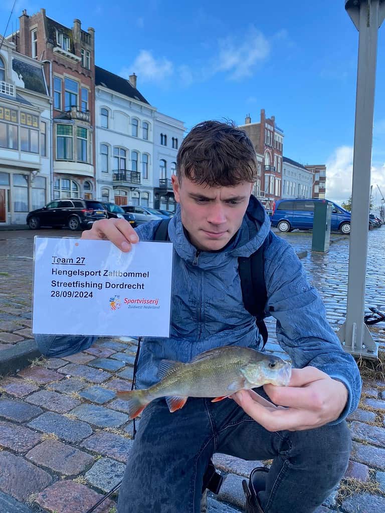 Een persoon in een blauw jasje houdt een vis vast en een bord met de tekst: "Team 27 Hengelsport Zaltbommel Streetfishing Dordrecht 28/09/2024" op een geplaveide straat met op de achtergrond historische panden, tijdens de NK Streetfishing kwalificatiewedstrijd.