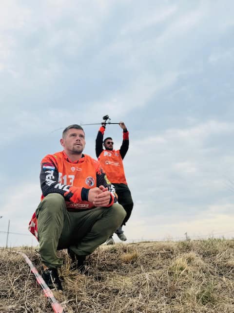 Twee mannen in oranje truien, waarschijnlijk bezig met een buitenactiviteit. Een hurkt op de voorgrond, terwijl de ander achterin staat en een voorwerp boven zijn hoofd houdt tegen een bewolkte lucht. Ze maken deel uit van het Nationaal Karperteam, met media-aandacht onder leiding van Cindy Horstman.