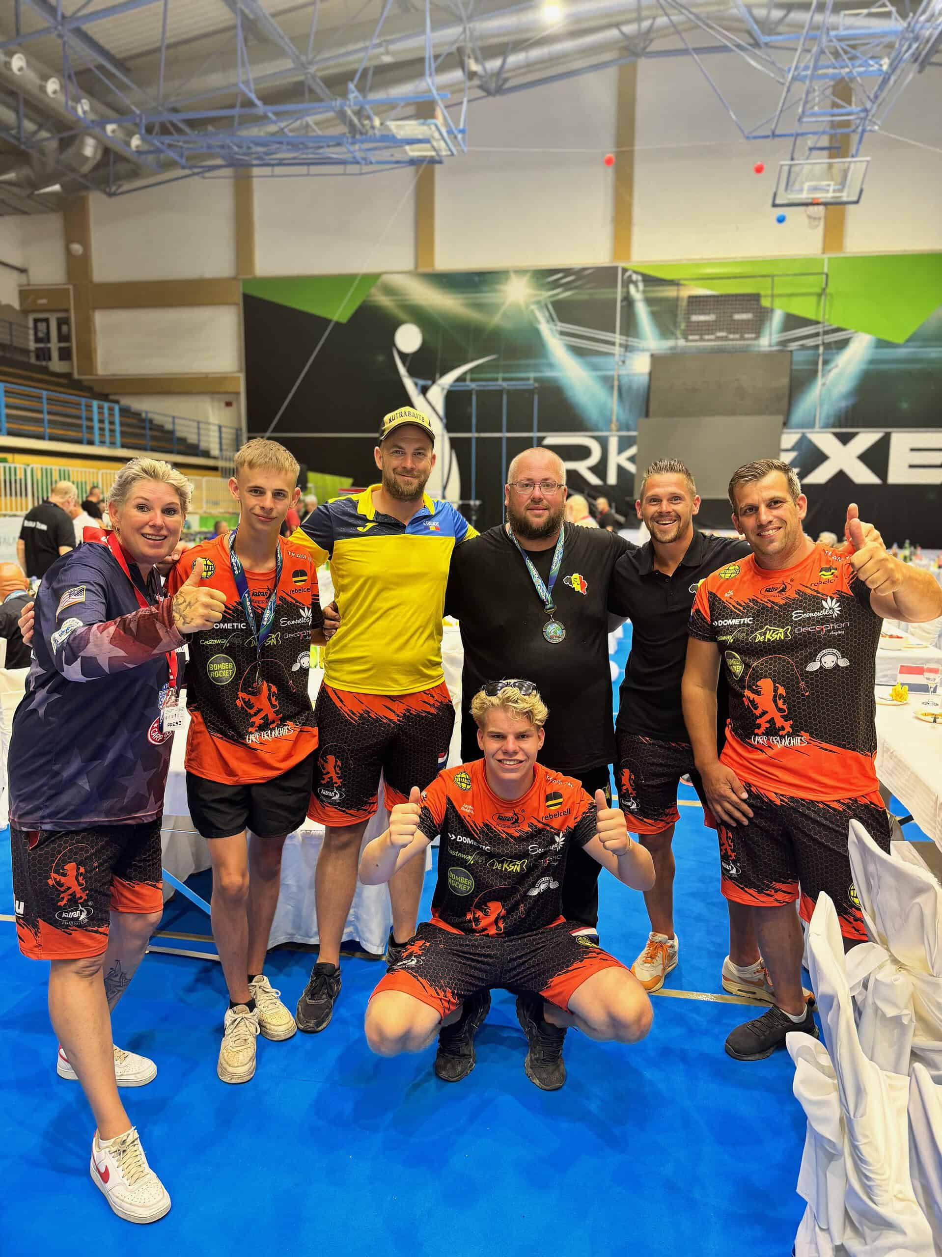 Een groep van zeven personen, waaronder NK's Media Manager Cindy Horstman, poseren samen op een blauwe vloer in een indoor sportfaciliteit. Ze dragen sportkleding, tonen trots hun medailles en glimlachen met hun duimen omhoog.