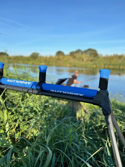 Een persoon vist bij een rivier, gedeeltelijk aan het zicht onttrokken door gras en een blauwe hengelhouder met het merk "Absolute" op de voorgrond, die doet denken aan scènes uit een verslag van het NK Dobbervissen door Jelle Zwijnenberg.