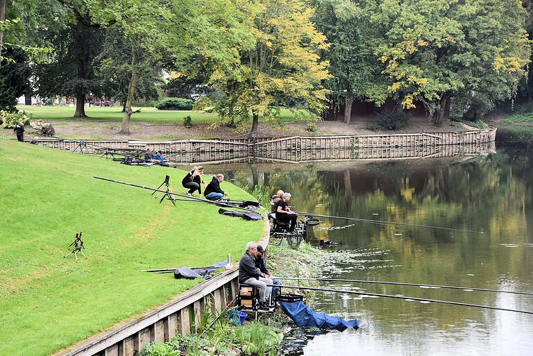 Mensen vissen langs een rivieroever met hengels die over het water uitsteken. Het tafereel bestaat uit groen gras, bomen en een kalme rivier die de omgeving weerspiegelt. Onder hen is Cindy Horstman, die deelneemt aan het clubkampioenschap voor dobbervissen.