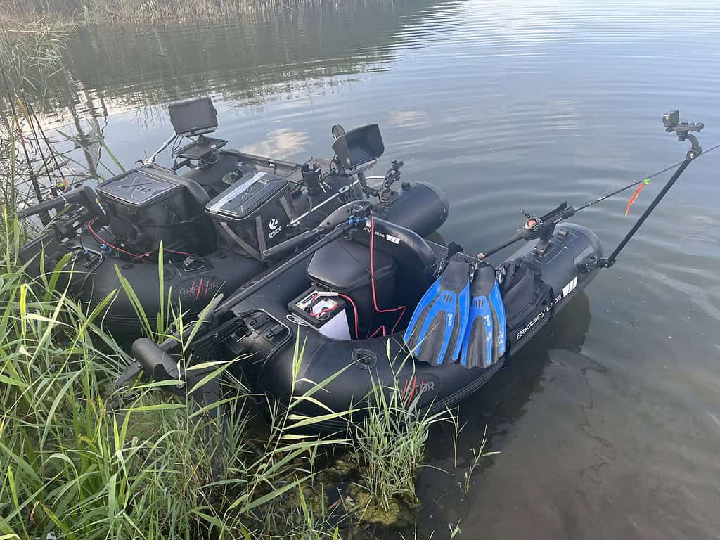 Twee kleine opblaasbare bellyboats uitgerust met motoren, visgerei en elektronische apparaten liggen aangemeerd aan de rand van een kalm waterlichaam, omringd door hoog gras. Ideaal voor een dagje Jeugddag met Sportvisserij Zuidwest Nederland.