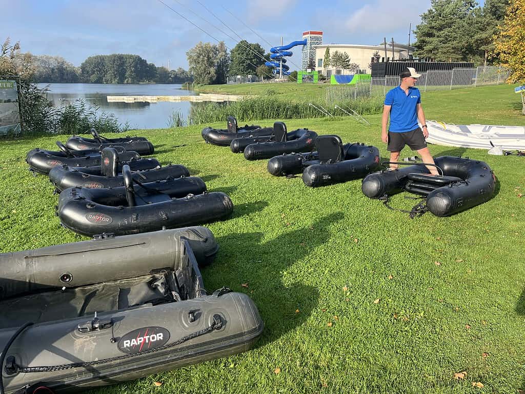 Een man staat op het gras naast een aantal opblaasbare bellyboats bij een meer tijdens de Jeugddag van Sportvisserij Zuidwest Nederland. Op de achtergrond zijn bomen, een gebouw en een waterglijbaan zichtbaar.