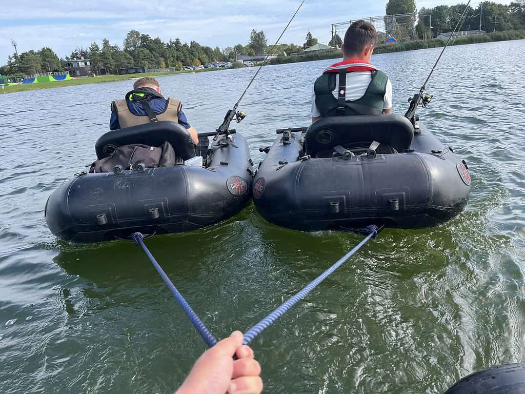 Twee mensen in bellyboats worden voortgetrokken door iemand die een touw vasthoudt op een meer met gebouwen en bomen op de achtergrond, wat doet denken aan een Jeugddag-evenement.