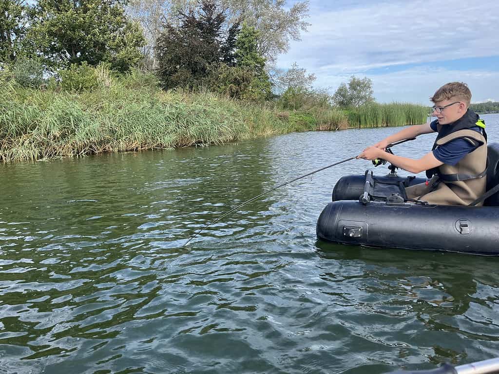 Een persoon in een kleine opblaasbare bellyboat vist in een kalme, groenkleurige rivier met hoog gras en bomen op een gedeeltelijk bewolkte Jeugddag. Hij geniet van de serene schoonheid van het water als onderdeel van Sportvisserij Zuidwest Nederland.