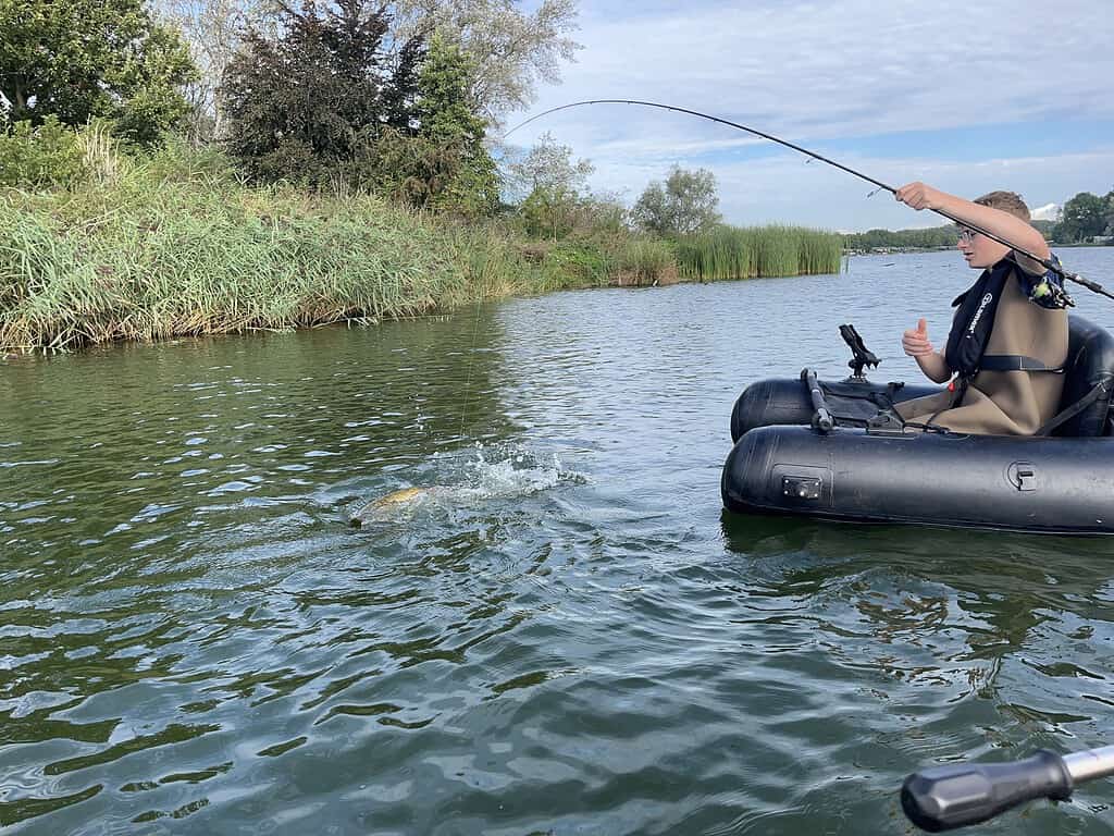 Een persoon in een zwarte opblaasbare bellyboat haalt een vis uit het water. De scène speelt zich af in een kalme, groene, natuurlijke waterweg met riet en bomen op de achtergrond, wat doet denken aan de serene Jeugddag-uitjes van Sportvisserij Zuidwest Nederland.