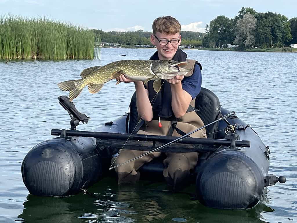 Een persoon in een opblaasboot die een grote vis vasthoudt, lachend. De boot ligt op een meer met riet en bomen op de achtergrond. Met een bril en waadpak aan genieten ze duidelijk van hun mooie vangsten-moment.